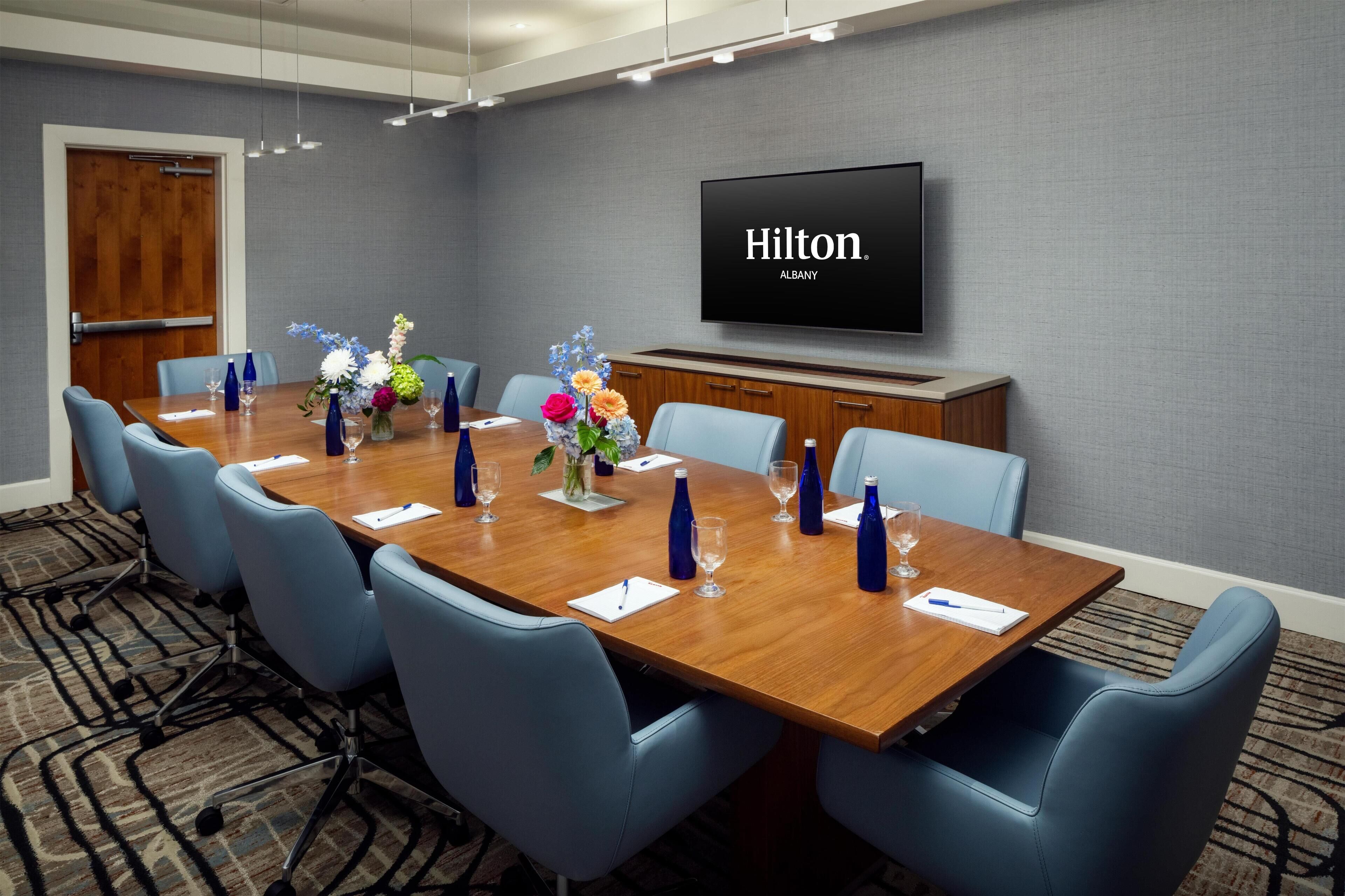Un salón de conferencias con una larga mesa, sillas azules, un televisor mostrando 'Hilton Albany' y centros de mesa con flores.