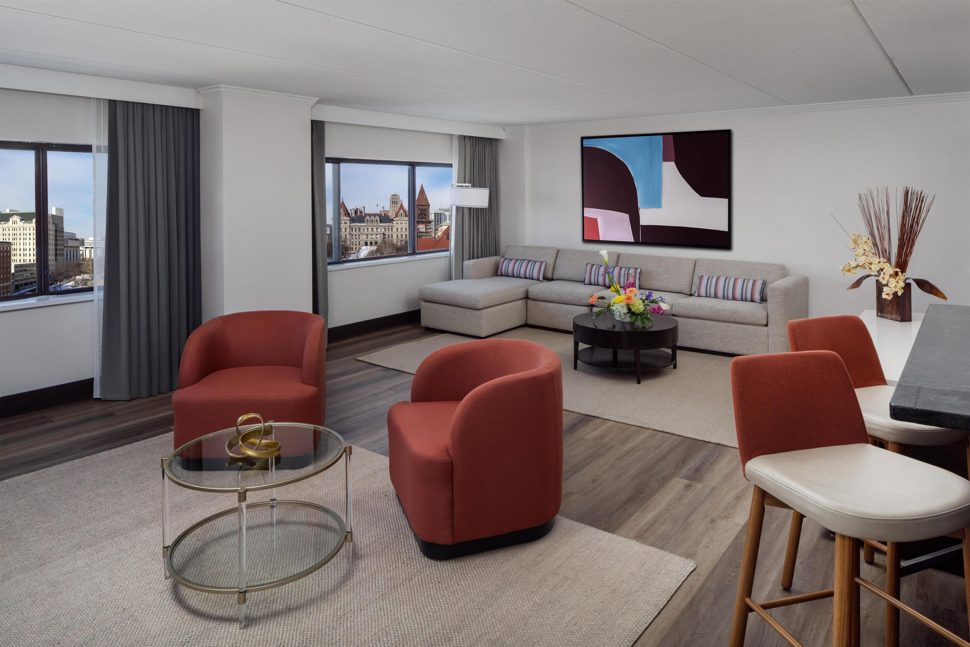 A modern hotel room with a sectional sofa, red armchairs, a glass coffee table, and a dining area.