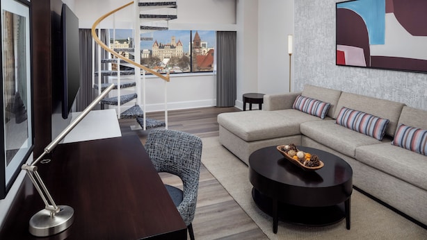 Una habitación de hotel moderna con un sofá, una mesa de centro, un escritorio con lámpara, vistas a un paisaje urbano a través de amplios ventanales y una escalera.