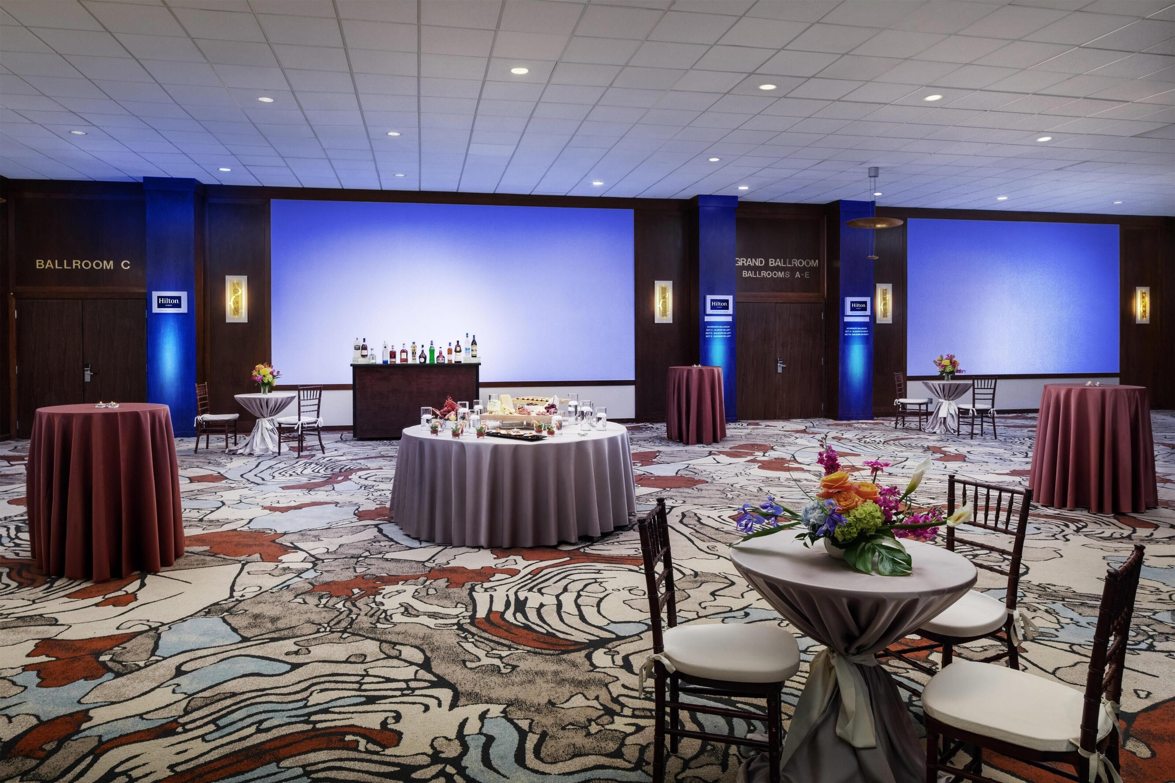 A spacious ballroom with a carpeted floor, round tables with chairs, and a stage with a blue backdrop.