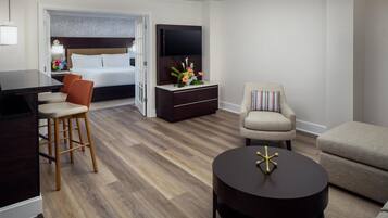 A modern hotel room with a wooden floor, a bed with white linens, a TV on a stand, a desk with a chair, and a sofa.