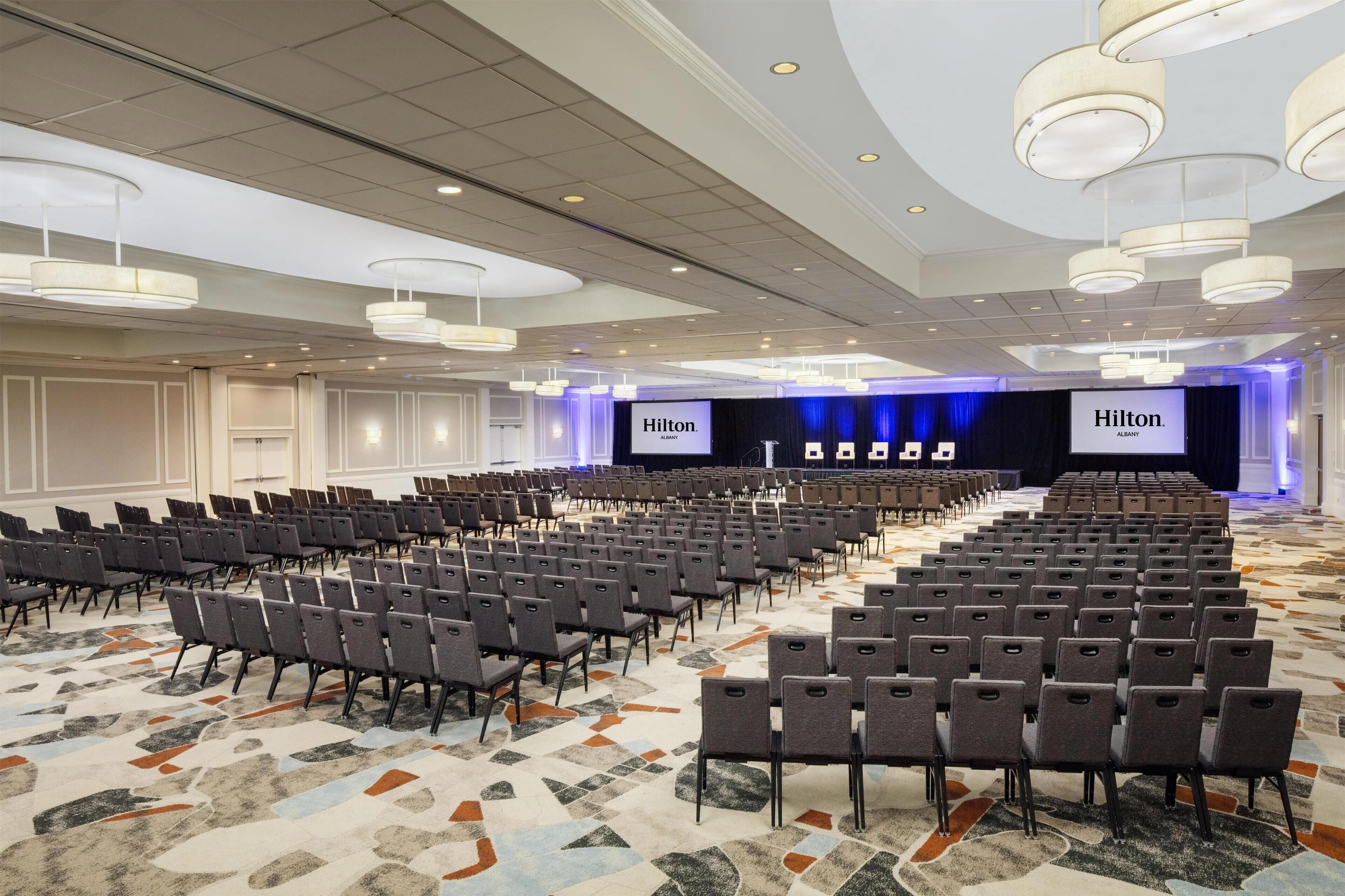 A large conference room with rows of chairs, a stage with a 'Hilton' banner, and a patterned carpet.