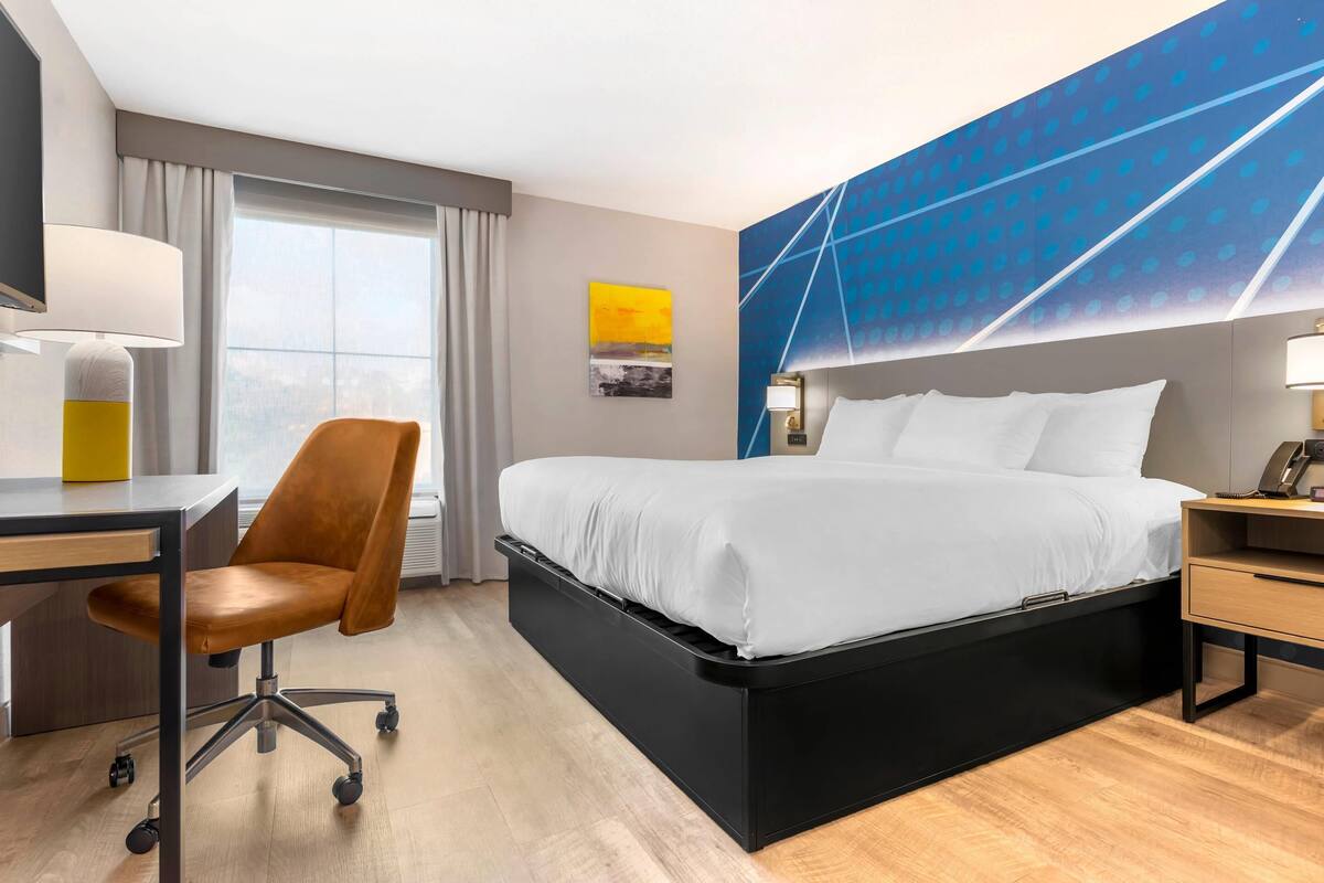 A modern hotel room with a bed, desk, chair, and large windows.