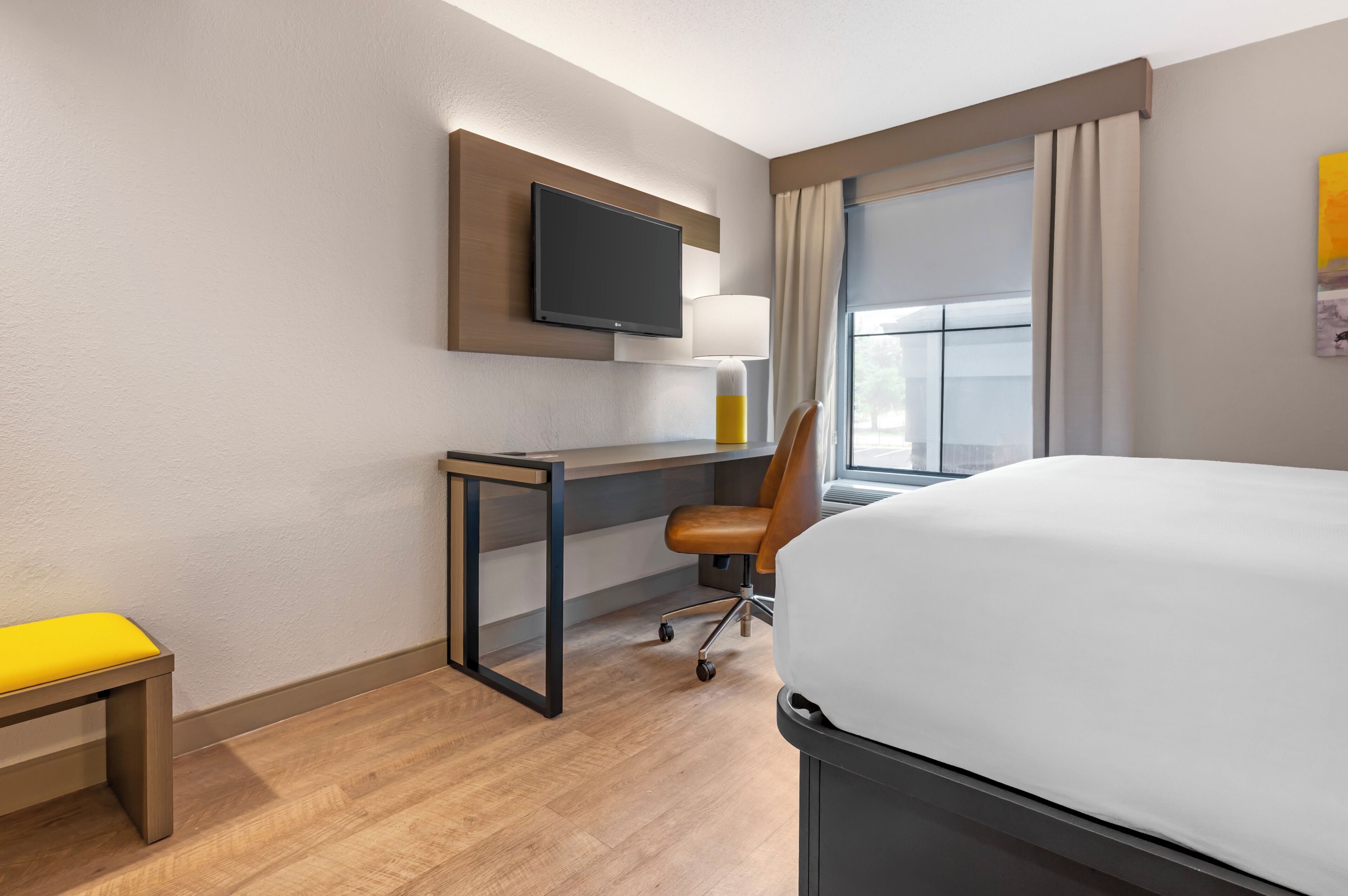 A hotel room with a bed, desk, chair, TV, window, and lamp.