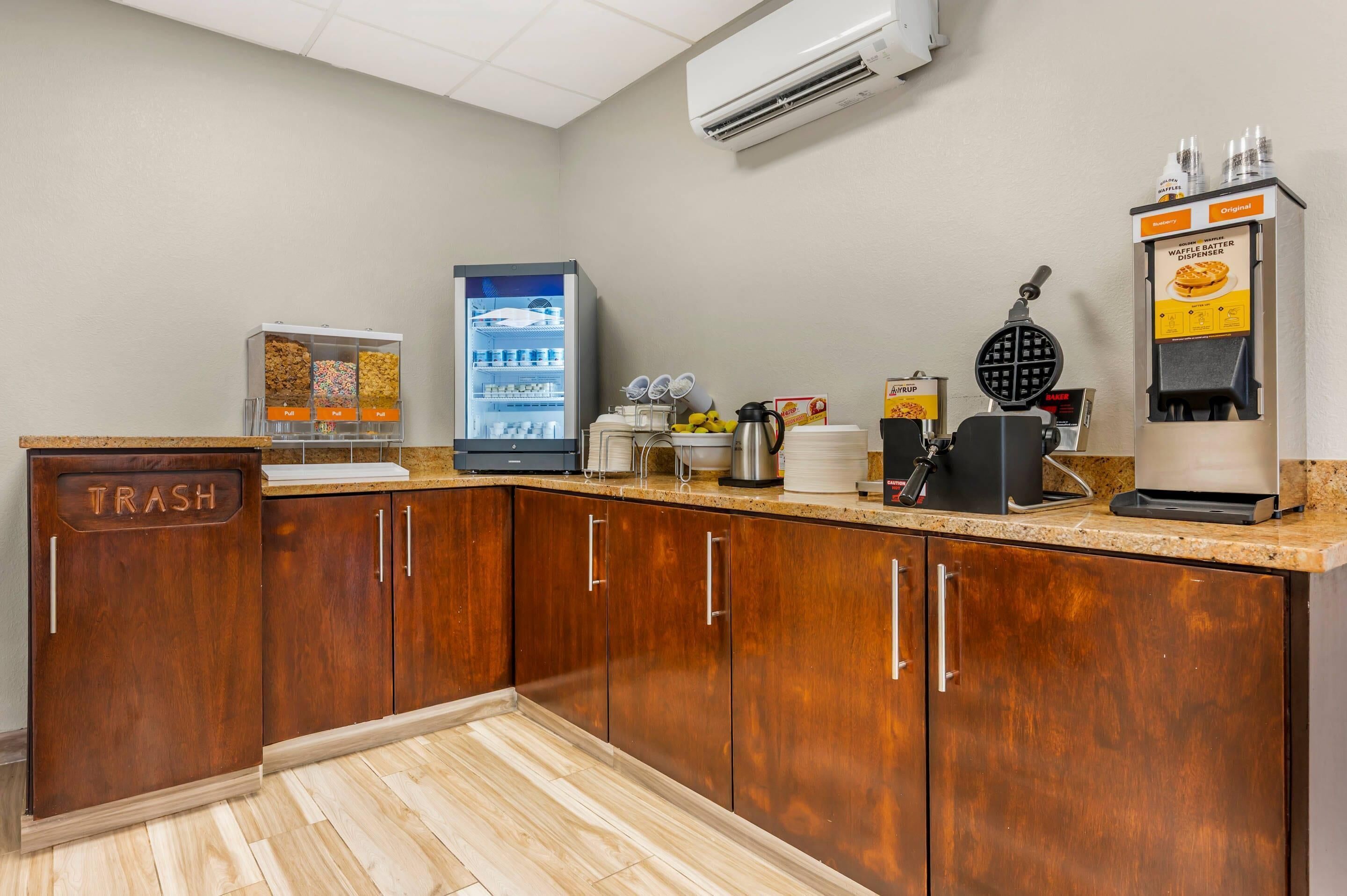 A kitchen area with a trash bin, a waffle batter dispenser, a waffle maker, and a refrigerator.