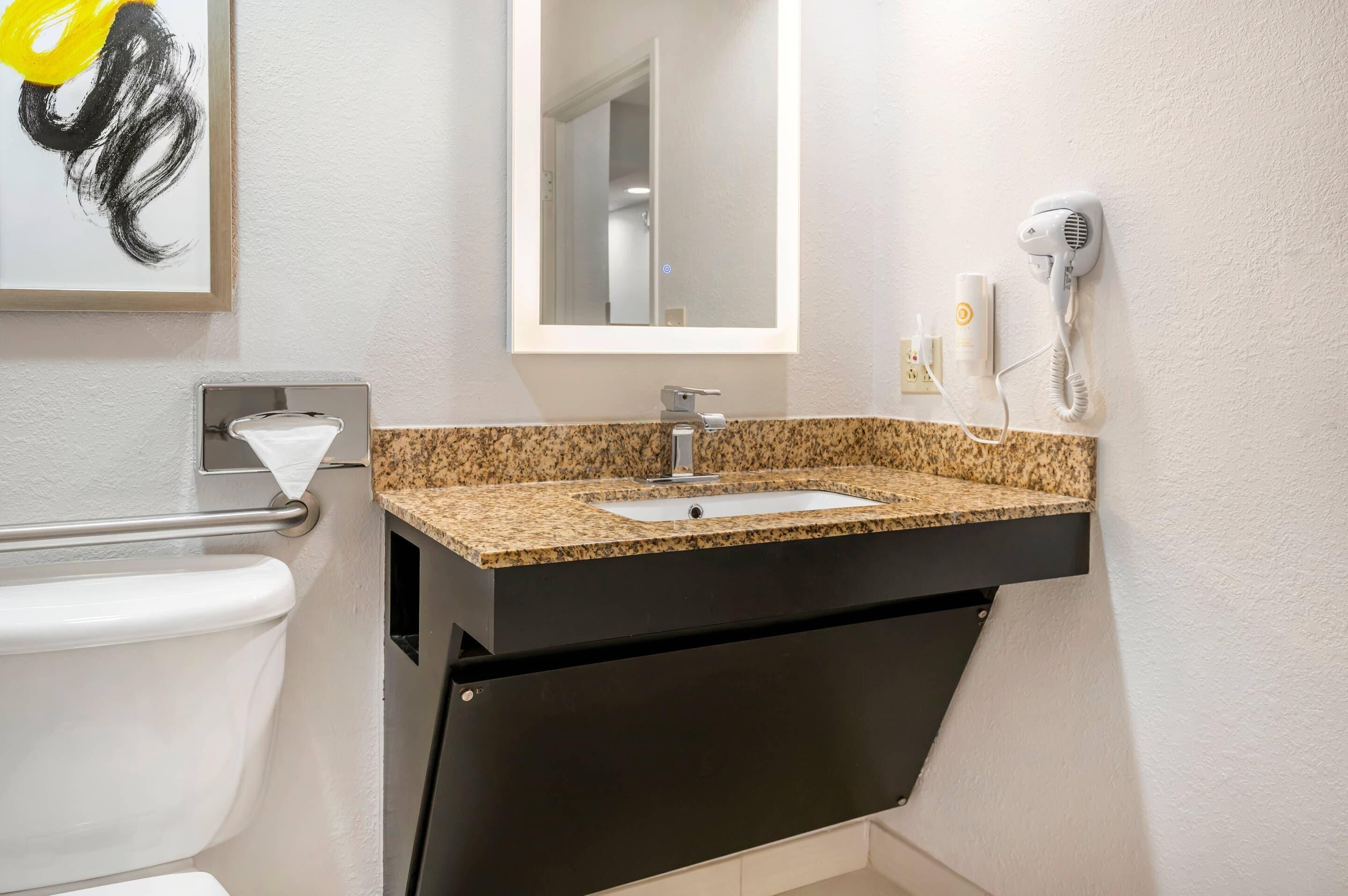 Un baño con lavamanos de granito, espejo, secador de pelo y dispensador de toallas.