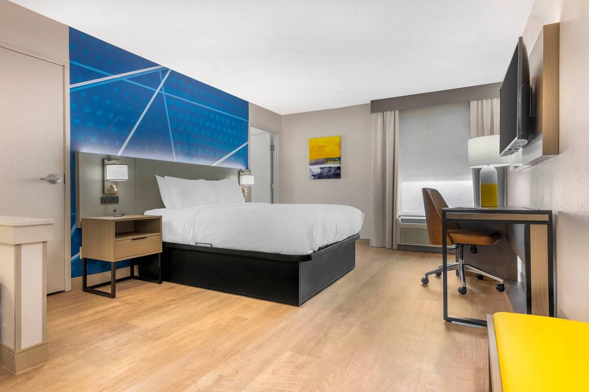 Quarto de hotel com cama, mesa de cabeceira, escrivaninha, cadeira e televisão presa na parede.