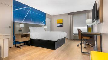 Ein Hotelzimmer mit Bett, Nachttisch, Schreibtisch, Sessel und einem an der Wand befestigten Fernseher.