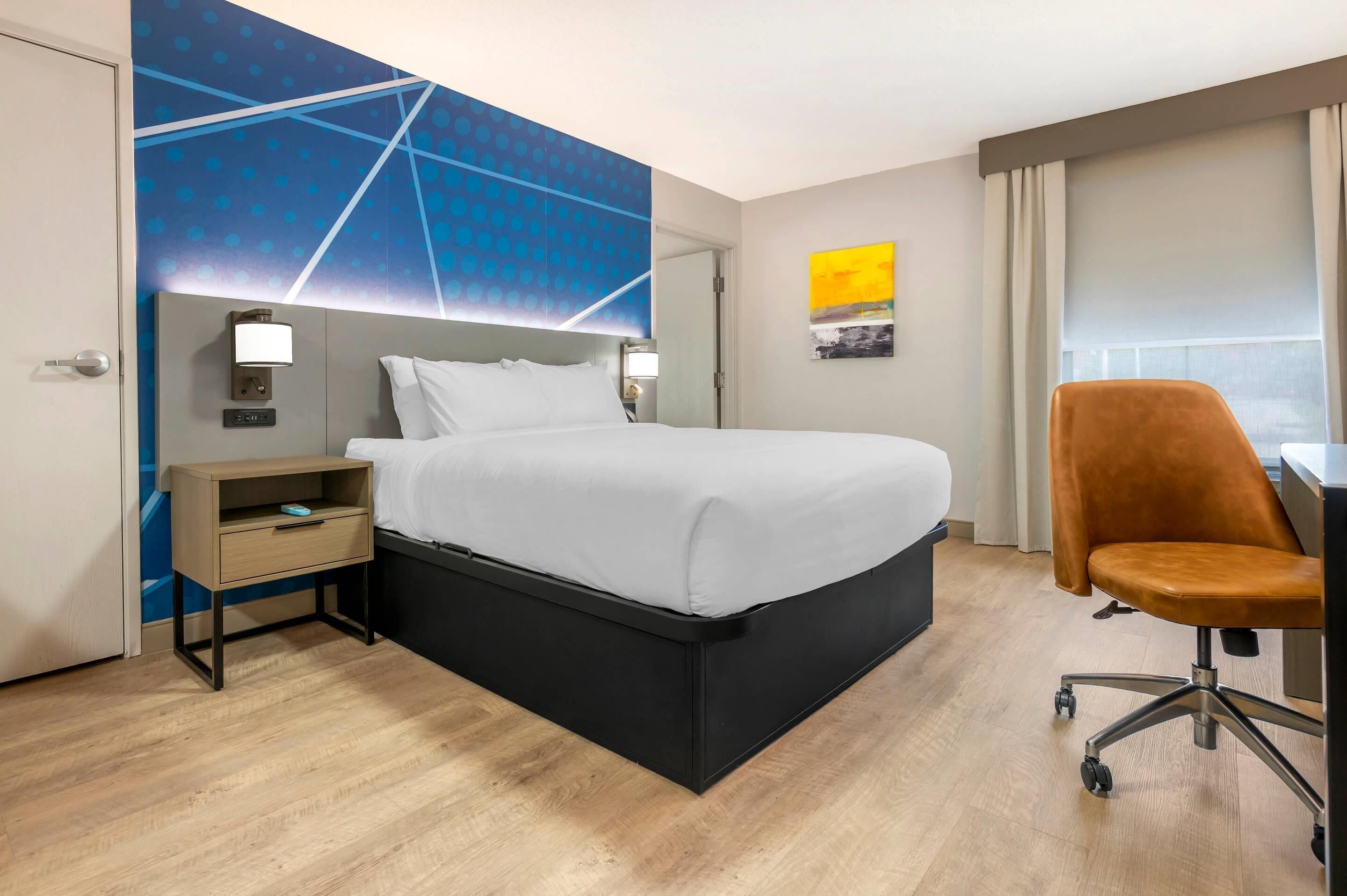 Ein Hotelzimmer mit Bett, Schreibtisch, Stuhl, Nachttisch, Wanduhr und einem Fenster mit Vorhängen.