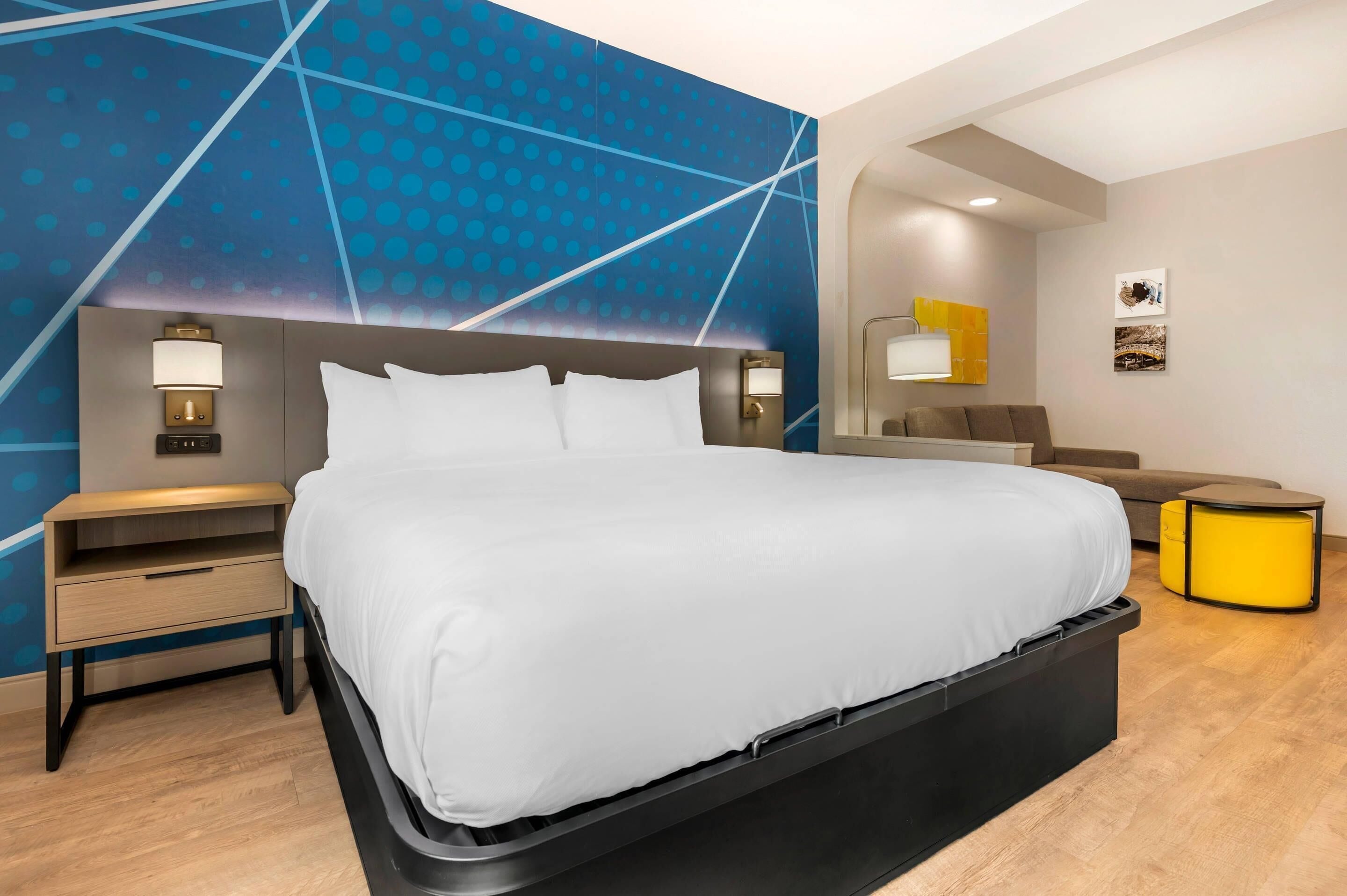 Une chambre d’hôtel moderne avec un grand lit, des tables de chevet, une petite table de nuit, un petit canapé, une table d’appoint jaune et un mur orné d’un motif géométrique.