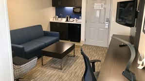 A compact living space with a sofa, a coffee table, a desk with a chair, a microwave, a refrigerator, and a television.