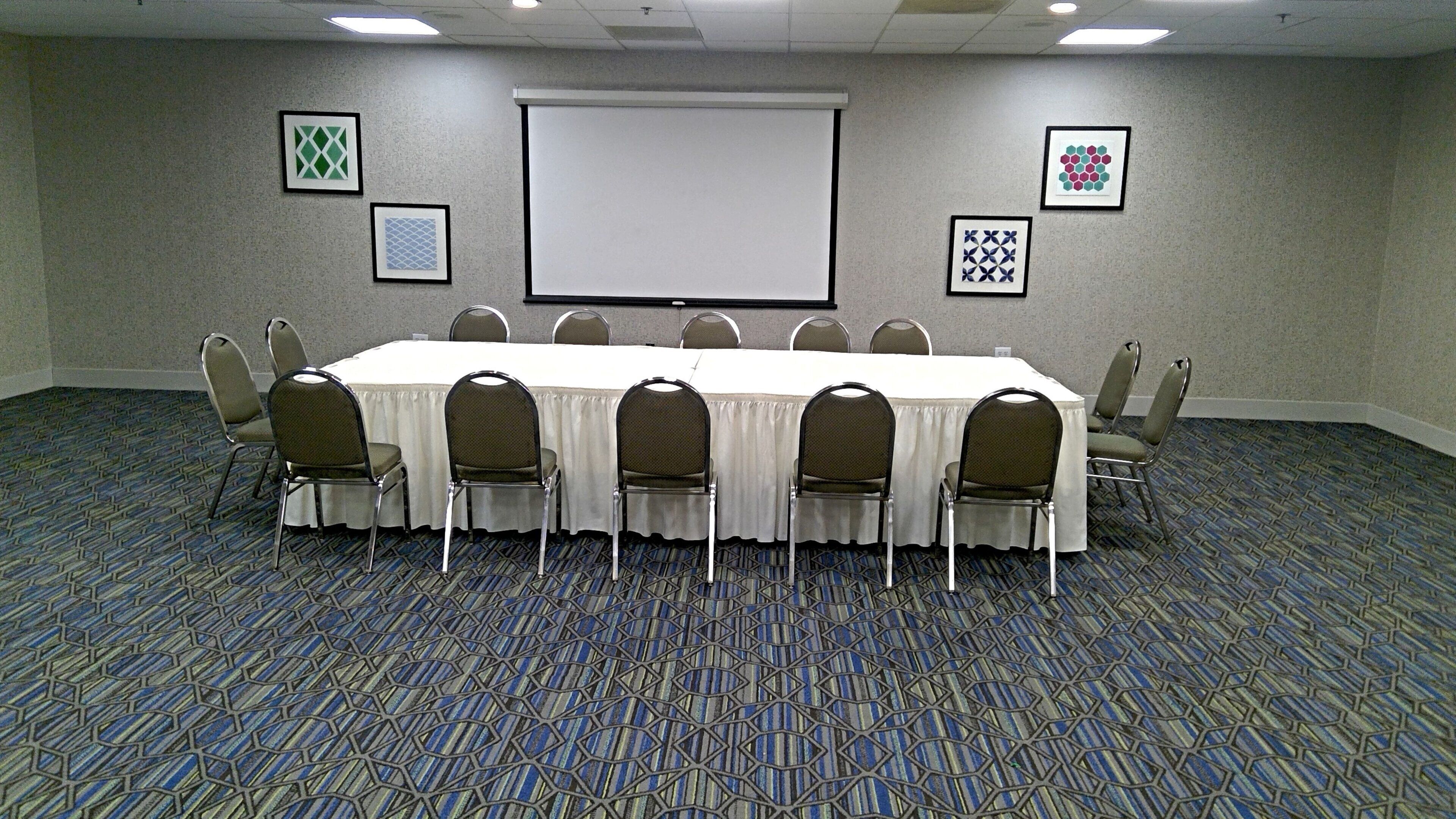 A conference room with a long table, chairs, a projector screen, and framed artwork on the walls.