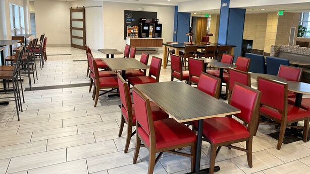 A restaurant with multiple tables and red chairs, a coffee bar, and a seating area with blue and brown chairs.