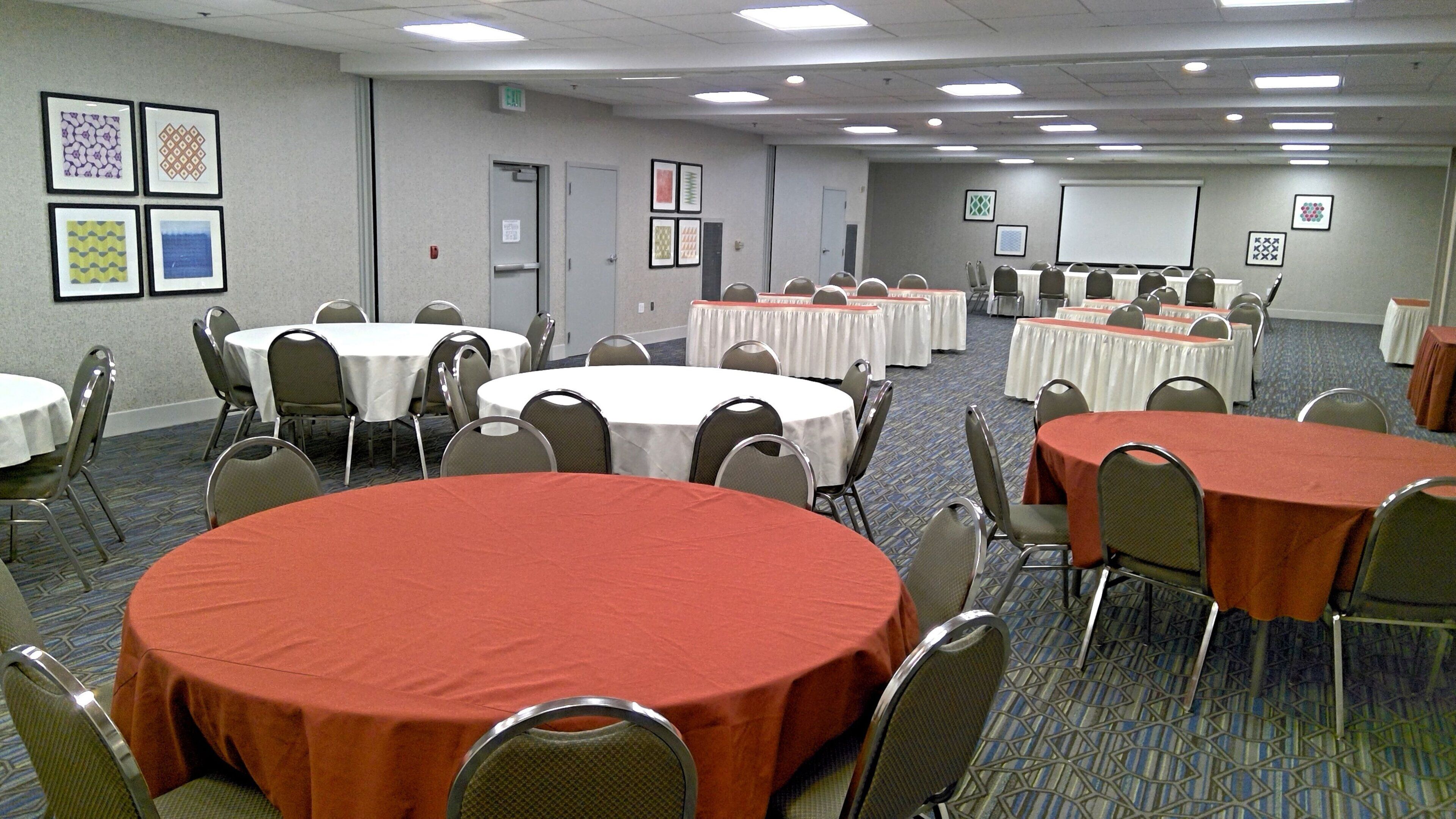 A conference room with multiple tables and chairs, a projector screen, and framed artwork on the walls.