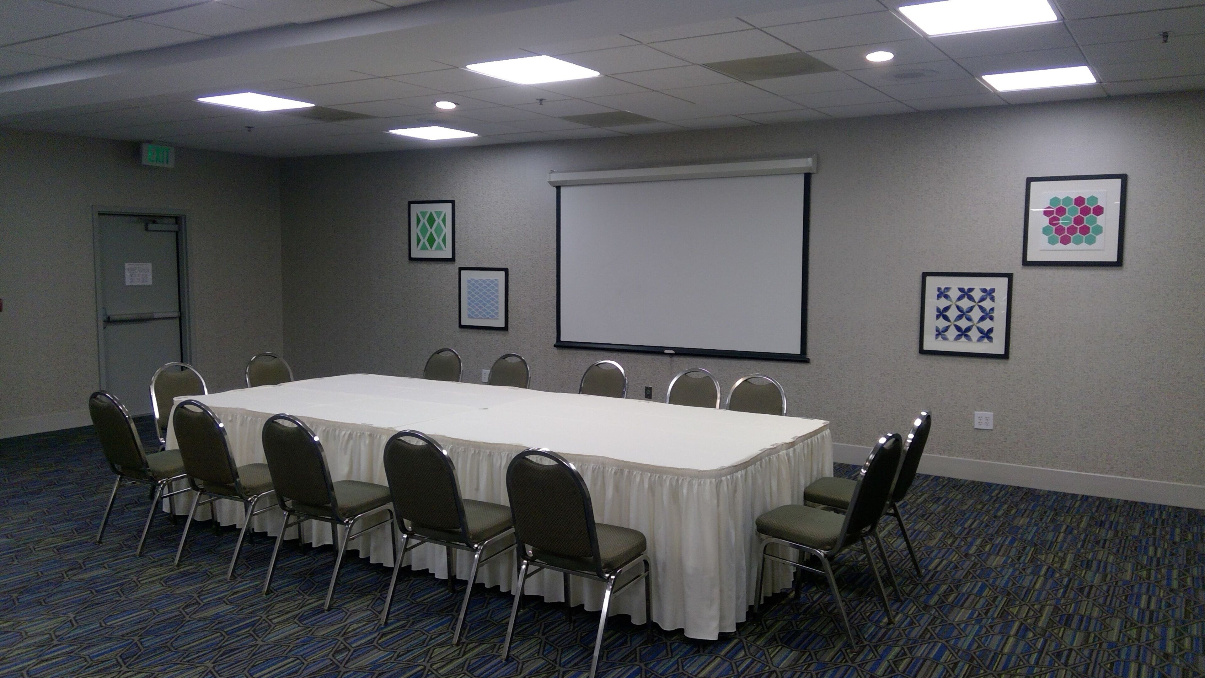 A conference room with a long table, chairs, a projector screen, and framed artwork on the walls.