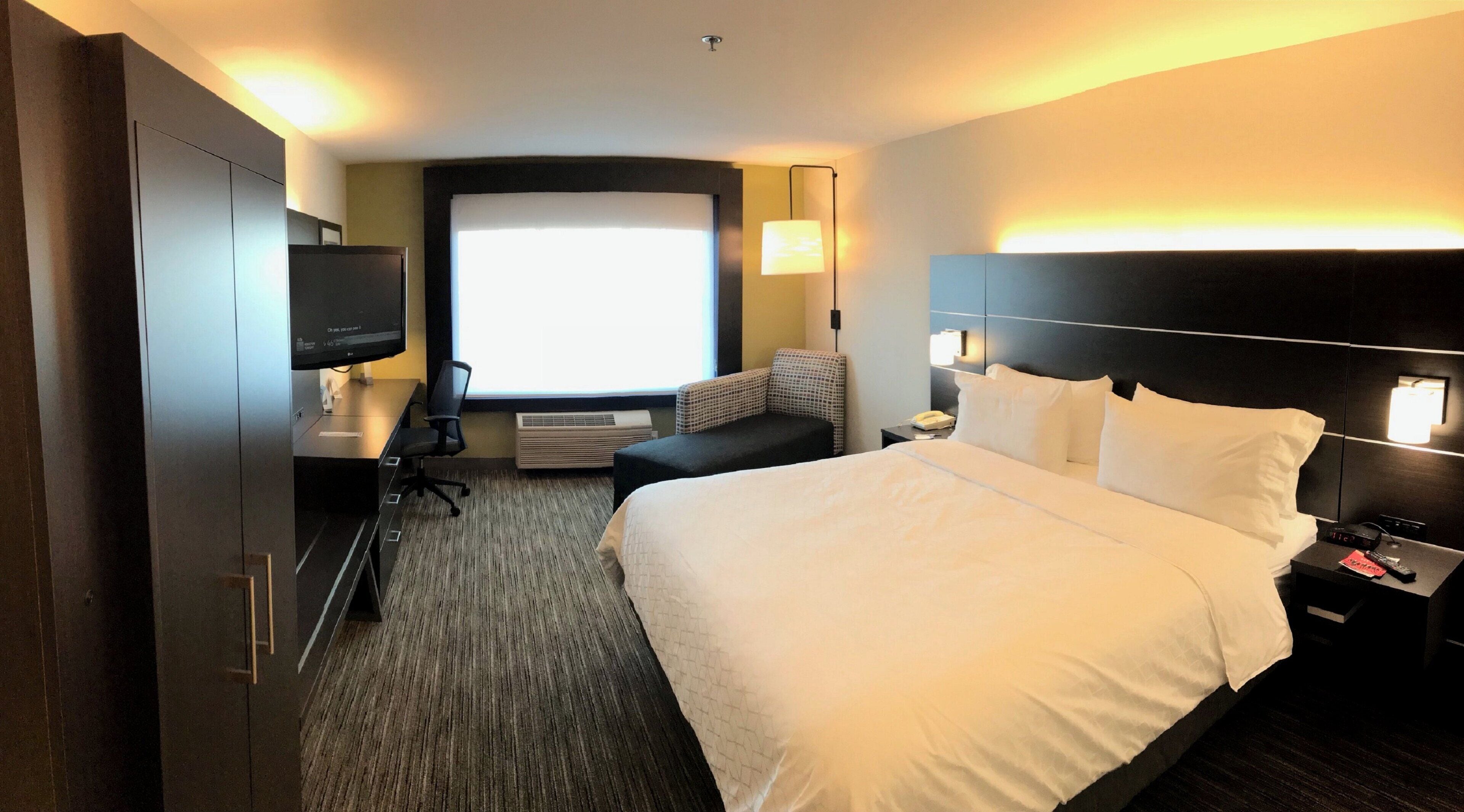 A hotel room with a large bed, a desk with a computer, a chair, a sofa, a TV, and a window with a view.