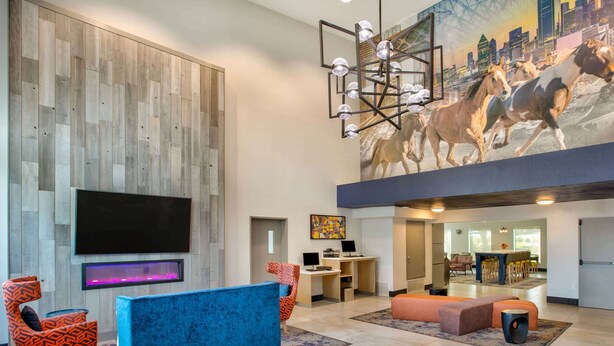 A modern lobby with a large flat-screen TV, a blue sofa, and a cityscape mural. - La Quinta Inn & Suites by Wyndham Dallas - Las Colinas (Irving)