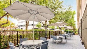 An outdoor patio area with round tables and chairs, large umbrellas, and a metal fence. - La Quinta Inn & Suites by Wyndham Dallas - Las Colinas (Irving)