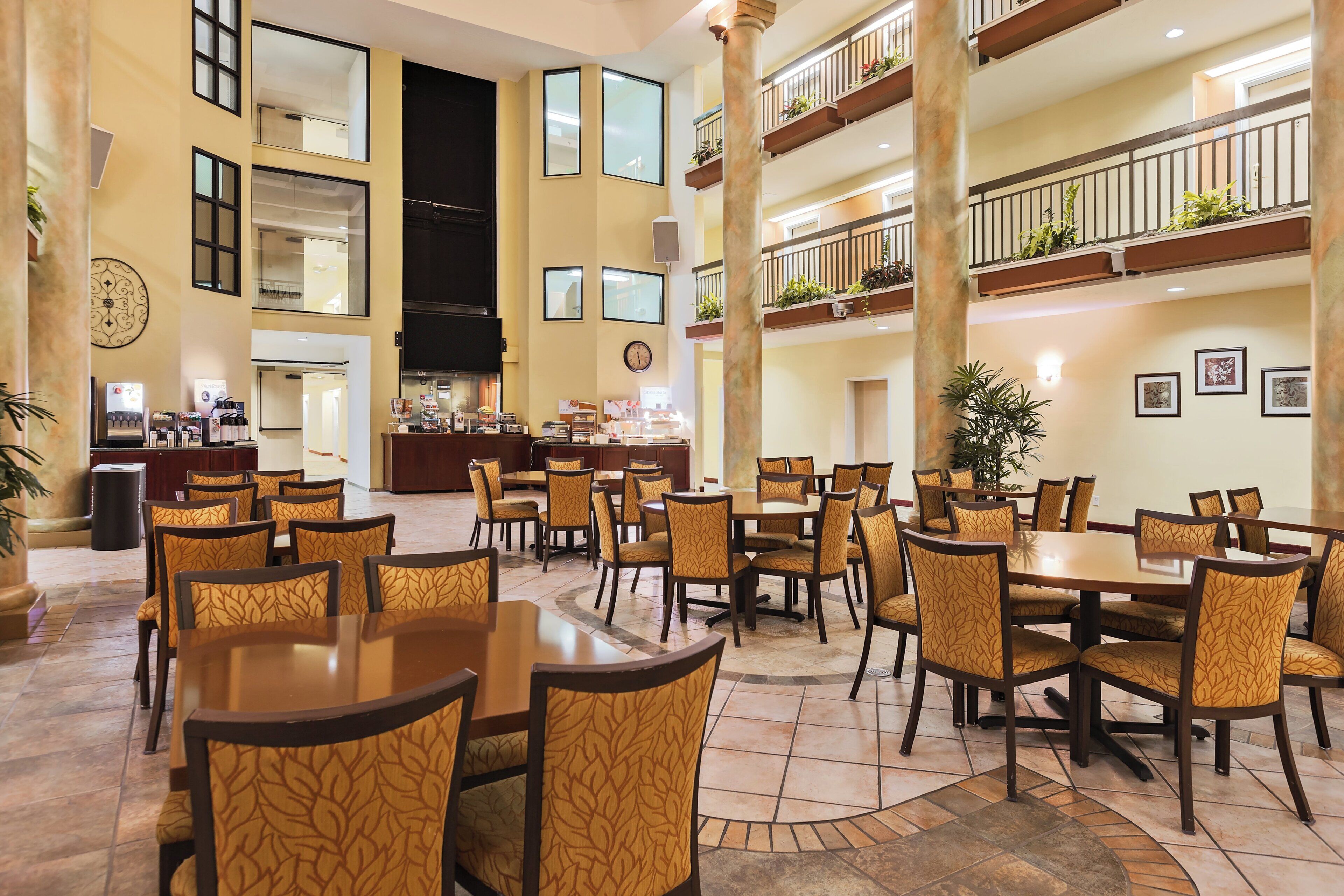 A spacious hotel lobby with multiple dining areas, a reception desk, and a large staircase connecting the floors.
