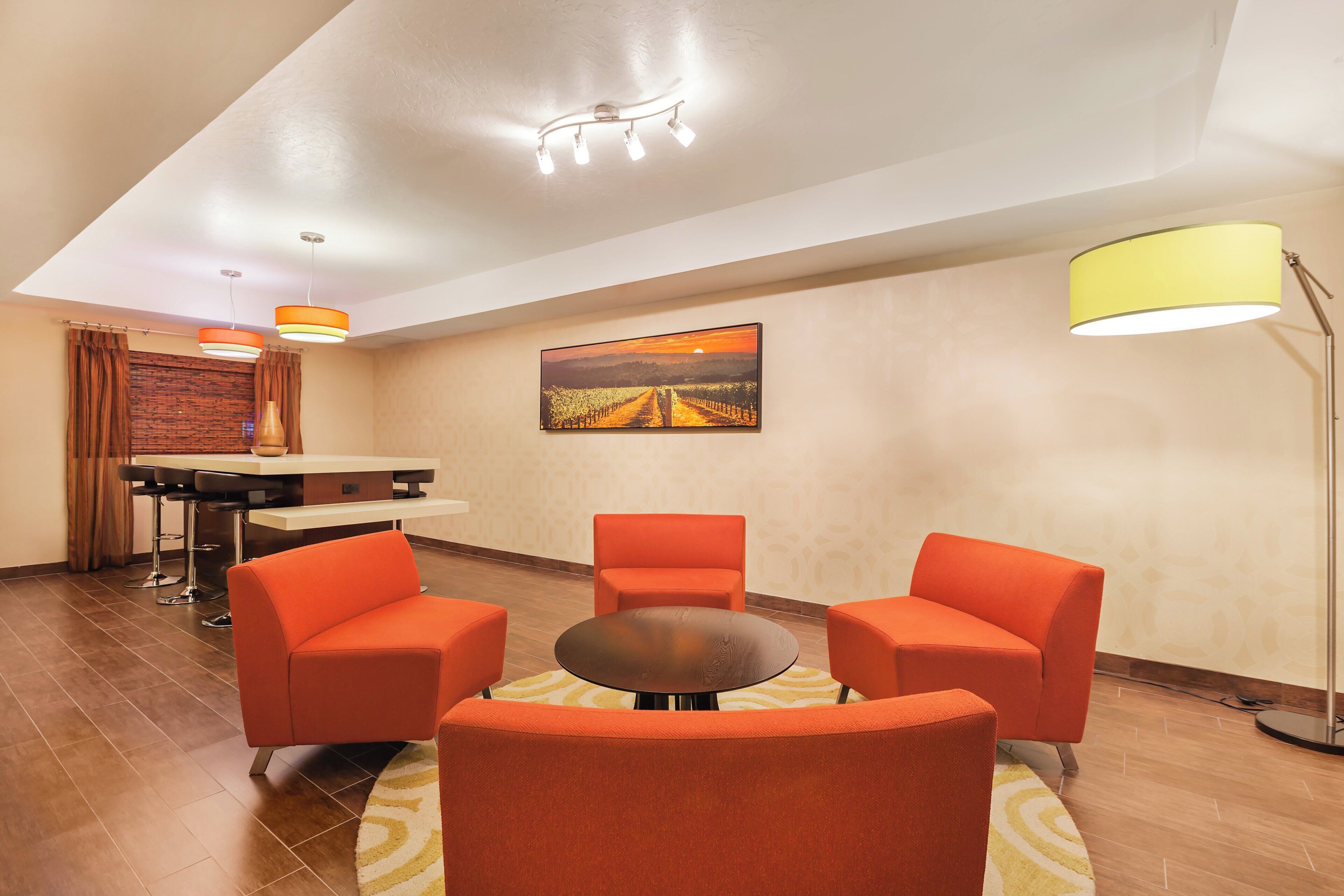 Habitación de hotel con comedor, dos sillas anaranjadas, una mesa redonda, un cuadro en la pared y una lámpara de pie.