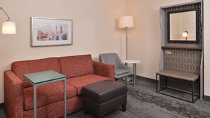 A hotel room with a red sofa, a grey armchair, a small round table, a bench, and a framed cityscape painting on the wall. - Holiday Inn Hotel & Suites Raleigh / Cary by IHG (Cary)