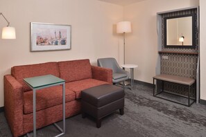 A hotel room with a red sofa, a grey armchair, a small round table, a bench, and a framed cityscape painting on the wall. - Holiday Inn Hotel & Suites Raleigh / Cary by IHG (Cary)