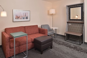 Habitación de hotel con un sofá rojo, un sillón gris, una mesita redonda pequeña, un banco y un cuadro con una vista urbana enmarcada en la pared.