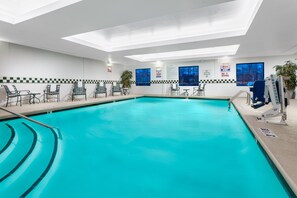 Una piscina interior con agua cristalina, rodeada de sillas y mesas de descanso, y paredes con señales de seguridad.