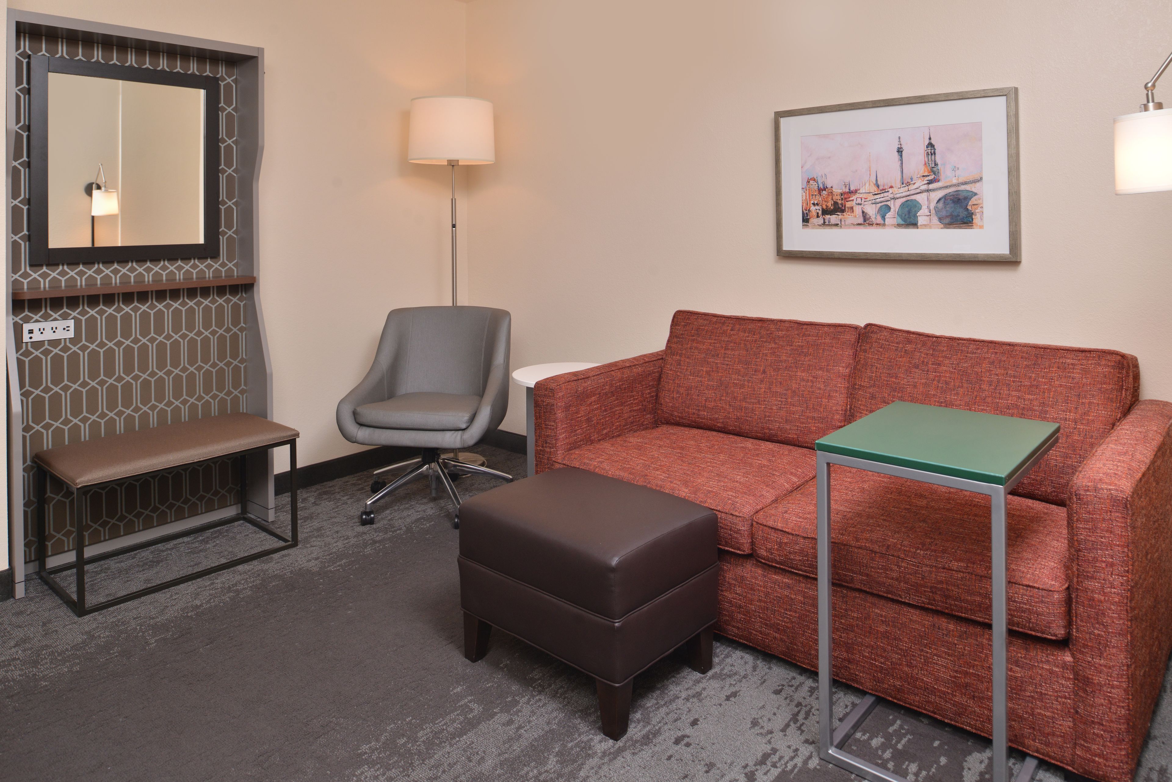 Une chambre d’hôtel avec un banc marron, un fauteuil de bureau gris, un canapé rouge, une petite table verte et un tableau représentant un paysage urbain, encadré et accroché au mur.