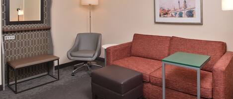 Une chambre d’hôtel avec un banc marron, un fauteuil de bureau gris, un canapé rouge, une petite table verte et un tableau représentant un paysage urbain, encadré et accroché au mur.