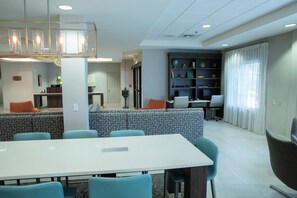 A modern dining area with a long table and chairs, a seating area with patterned cushions, and a bookshelf with various items. - Holiday Inn Hotel & Suites Raleigh / Cary by IHG (Cary)