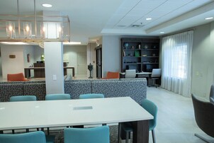 Un comedor moderno con una mesa larga y sillas, una zona de estar con cojines estampados, y una estantería con diversos objetos.