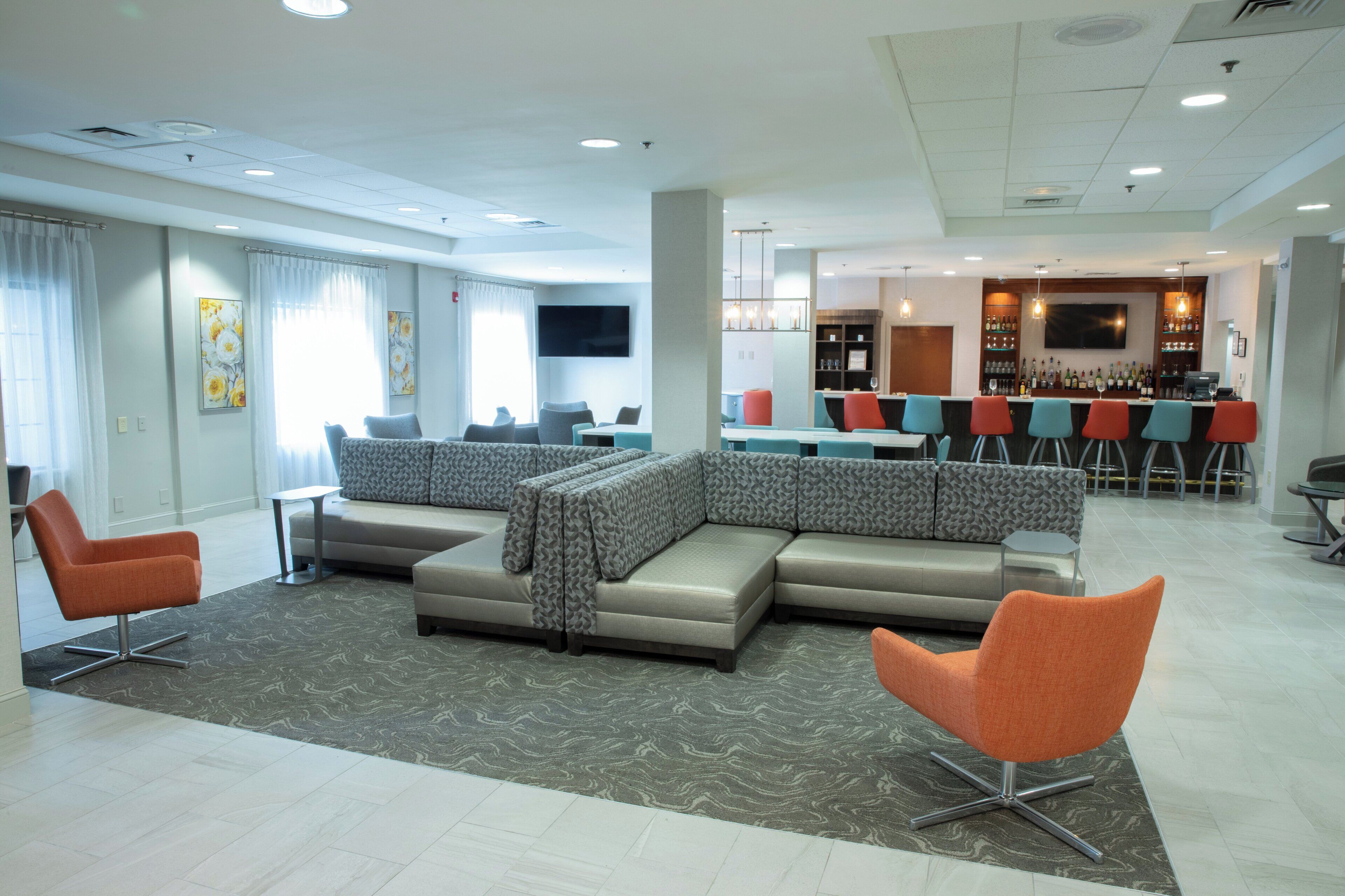A modern hotel lobby with a sectional sofa, armchairs, a bar area, and a flat-screen TV.