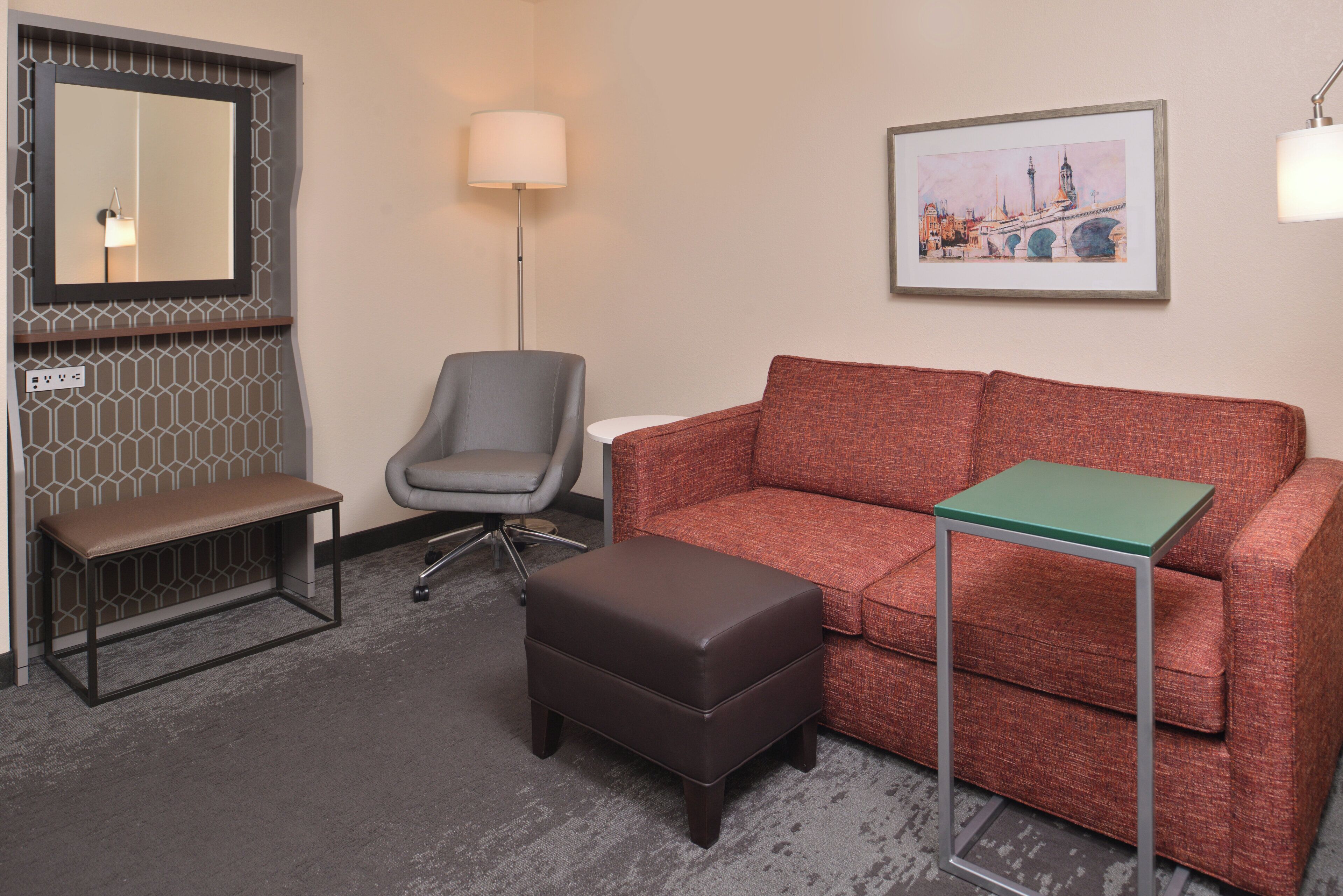 A hotel room with a grey office chair, a brown bench, a red sofa, a green side table, and a framed cityscape painting on the wall.