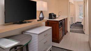 A compact hotel room with a flat-screen TV, a desk with a chair, a small kitchenette, and a bathroom visible through an open door. - Holiday Inn Hotel & Suites Raleigh / Cary by IHG (Cary)