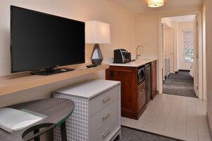 Habitación de hotel compacta con televisión de pantalla plana, escritorio con silla, kitchenette pequeña y baño visible a través de una puerta abierta.