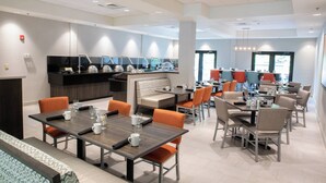 A modern dining area with multiple tables and chairs, a kitchen area with a counter and appliances, and a seating area with a bench. - Holiday Inn Hotel & Suites Raleigh / Cary by IHG (Cary)