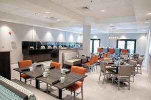 A modern dining area with multiple tables and chairs, a kitchen area with a counter and appliances, and a seating area with a bench. - Holiday Inn Hotel & Suites Raleigh / Cary by IHG (Cary)