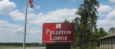 Un letrero para Pellston Lodge, un hotel independiente MagnuSon, con un asta de banderas y árboles de fondo.