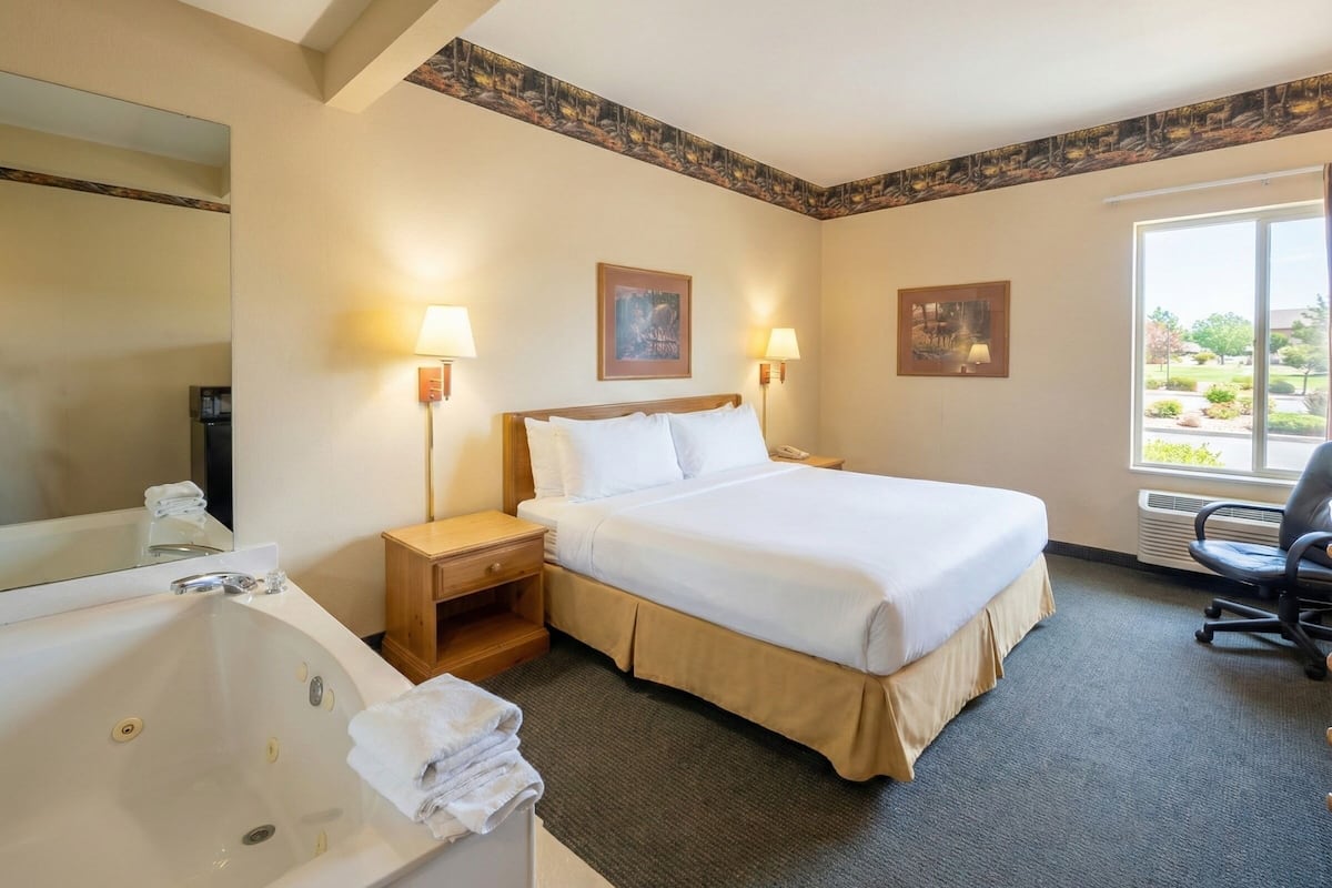 A hotel room with a large bed, a sitting area with a chair, a bathtub, and two wall-mounted lamps.