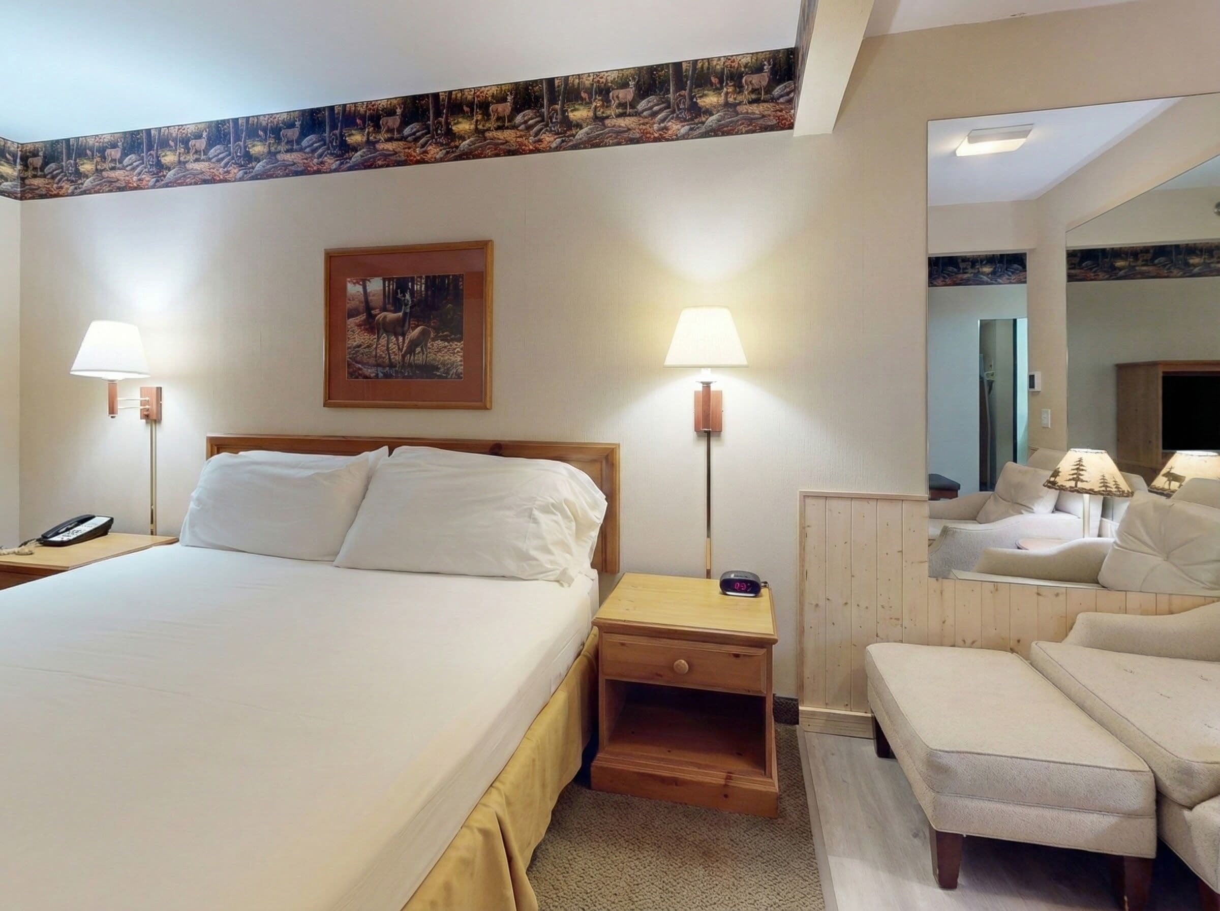 Habitación de hotel con una cama grande, dos mesitas de noche con lámparas, una mesita de madera, una pequeña mesa con un reloj, un espejo y un sofá con un reposapiés a juego.