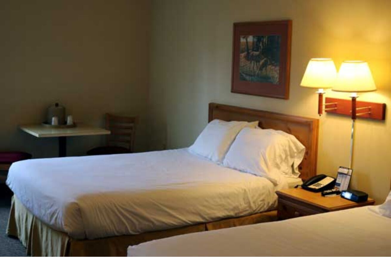 A hotel room with two beds, a bedside table with a lamp, a chair, and a painting on the wall.