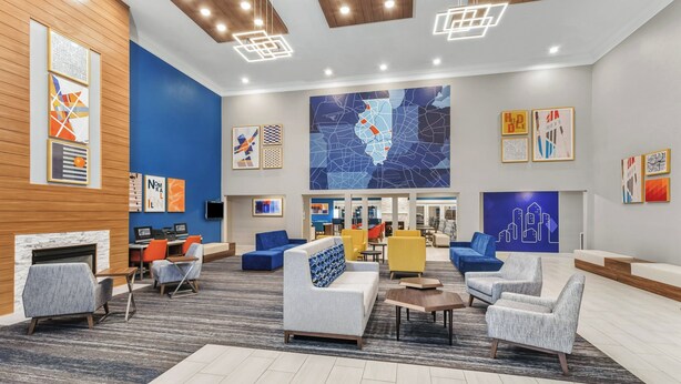 A modern lobby with seating areas, a fireplace, and a large map mural on the wall. - Holiday Inn Express & Suites Bloomington - Normal by IHG (Normal)