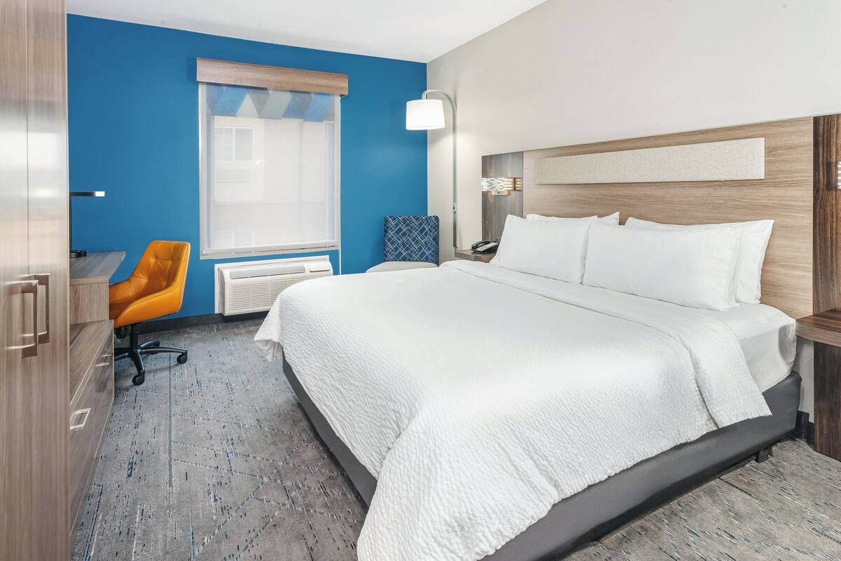 A hotel room with a large bed, a desk with a chair, a window with blinds, and a nightstand with a lamp.