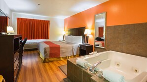 Hotelkamer met een bed, bureau, stoel, bureaulamp, nachtkastje, spiegel, badkuip en raam met gordijnen.