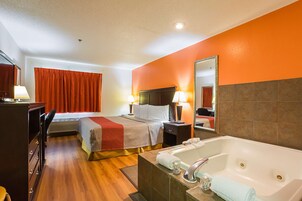Habitación de hotel con cama, escritorio, silla, lámpara de escritorio, mesita de noche, espejo, bañera y ventana con cortinas.