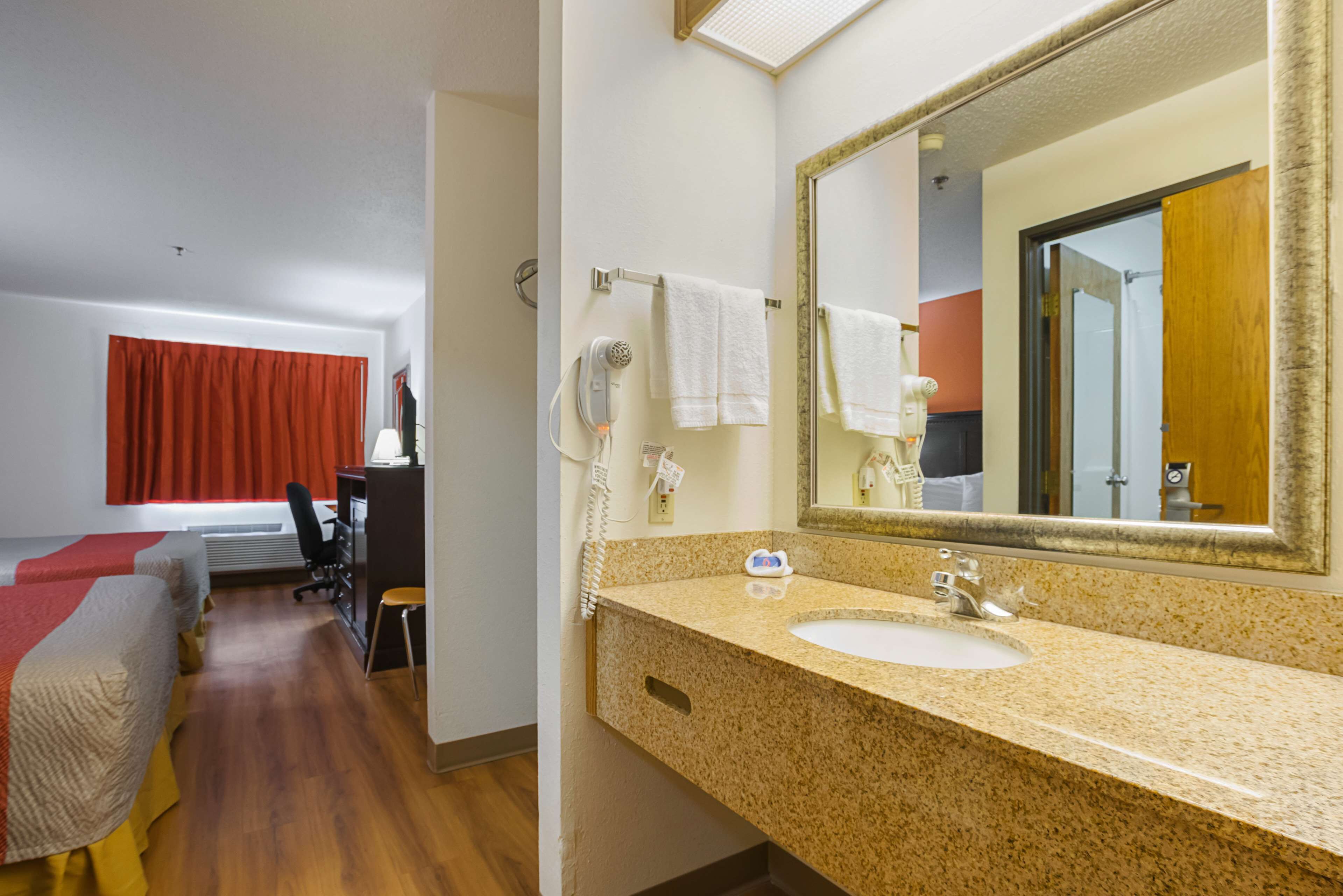 A hotel room with a bed, a desk, a chair, a bathroom with a sink and mirror, and a red curtain.