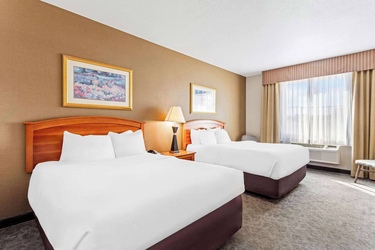 A hotel room with two beds, a wooden headboard, a nightstand with a lamp, a framed picture on the wall, and a large window with curtains.