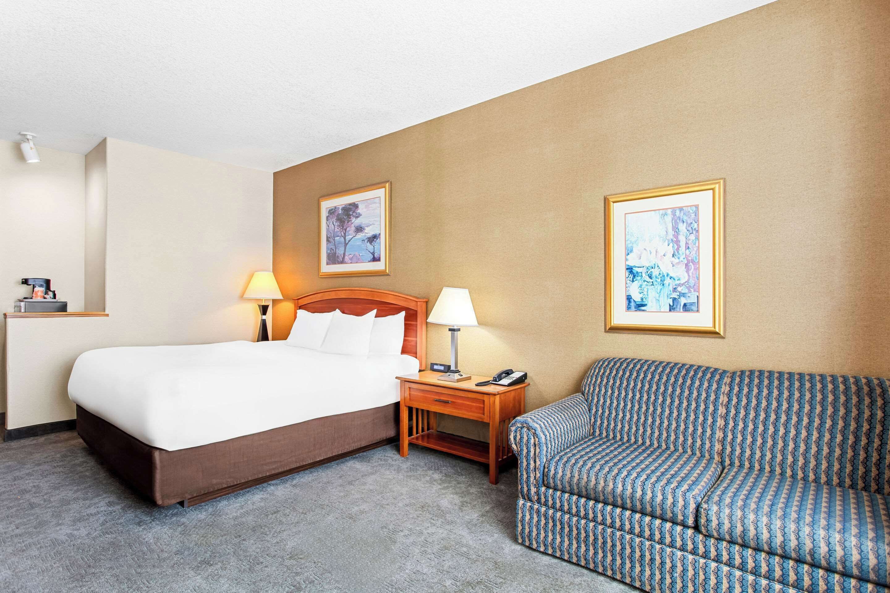 A hotel room with a bed, a nightstand, a lamp, a sofa, and two framed pictures on the wall.