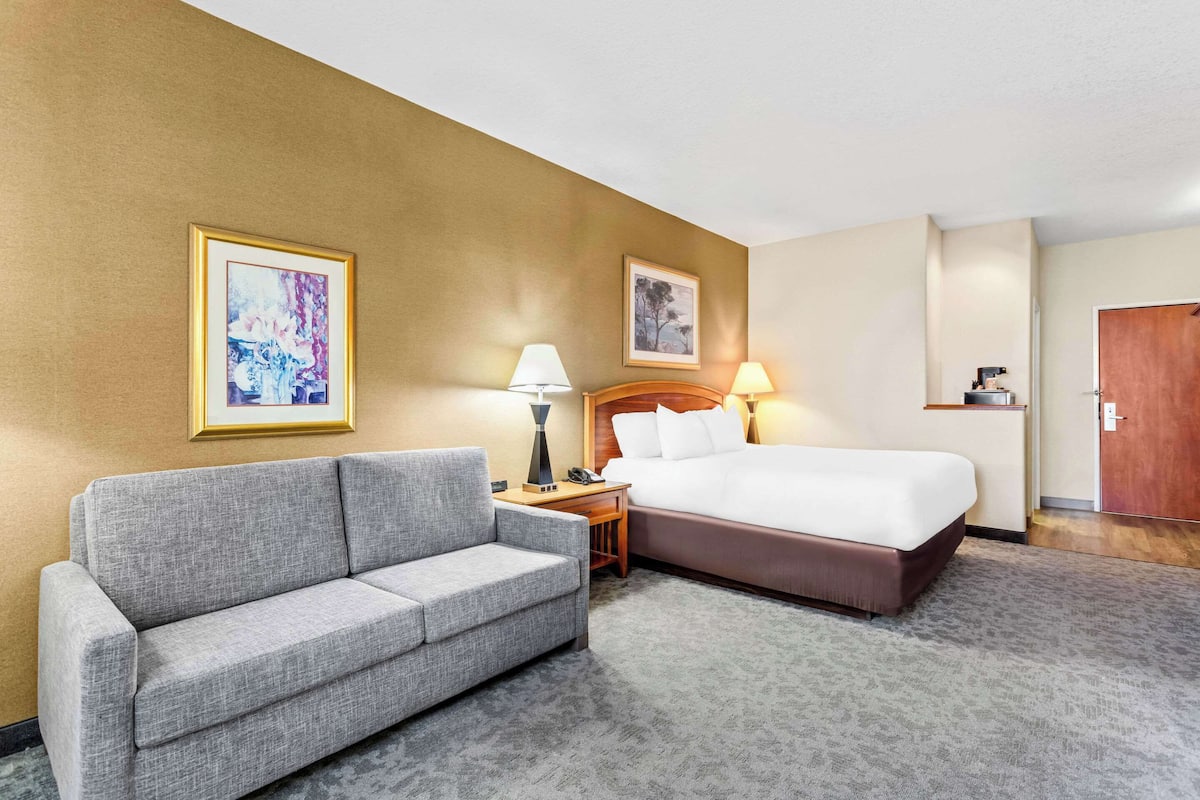 A hotel room with a gray sofa, a bed with white linens, two wall-mounted lamps, and framed artwork on the walls.
